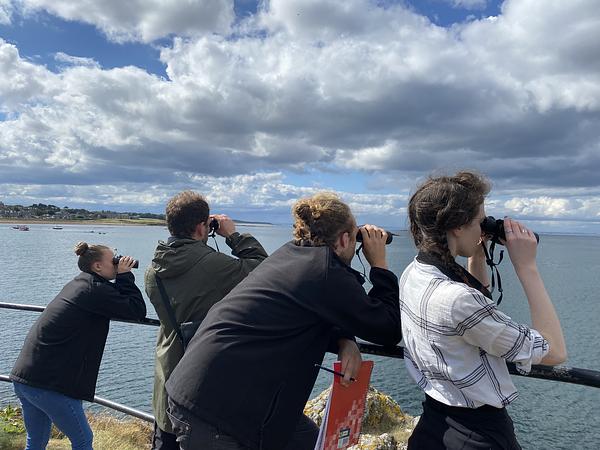 A group of 4 volunteers gaze out to sea through their binoculars on a bright day in North Berwick with scattered clouds.