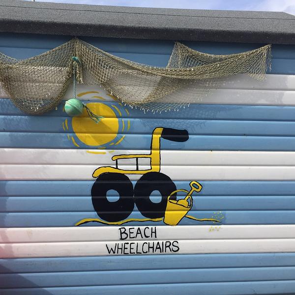 The beach wheelchair beach hut is painted with blue and white stripes and decorated with draping fishing nets and buoys. On the side of the hut, the Beach Wheelchair logo is painted: a bright yellow wheelchair with big black wheels upon a strip of sand and beside a yellow bucket and spade. A big yellow sun is painted above it, "BEACH WHEELCHAIRS" below. 