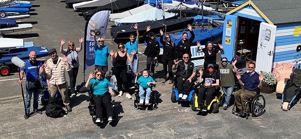 A group of 16 people smile and wave up at the camera from beside the blue and white striped Beach Wheelchair beach hut at North Berwick Harbour. The group includes a couple of film crew, the Beach Wheelchair team in bright blue t-shirts, three of the Scottish Seabird Centre team, and eight people from Euan's Guide including its ambassadors.