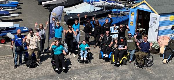 A group of 16 people smile and wave up at the camera from beside the blue and white striped Beach Wheelchair beach hut at North Berwick Harbour. The group includes a couple of film crew, the Beach Wheelchair team in bright blue t-shirts, three of the Scottish Seabird Centre team, and eight people from Euan's Guide including its ambassadors. A group of 16 people smile and wave up at the camera from beside the blue and white striped Beach Wheelchair beach hut at North Berwick Harbour. The group includes a couple of film crew, the Beach Wheelchair team in bright blue t-shirts, three of the Scottish Seabird Centre team, and eight people from Euan's Guide including its ambassadors.