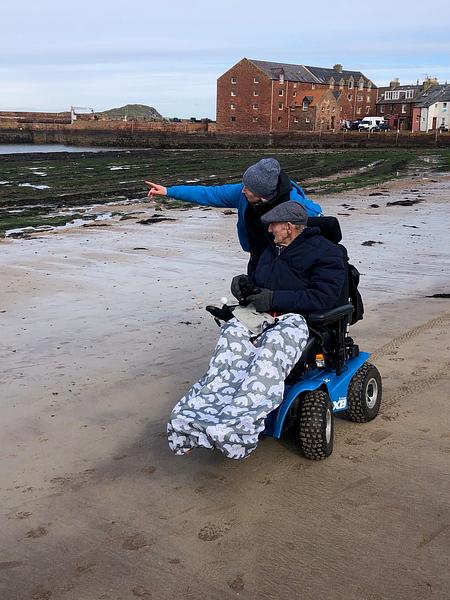 A volunteer from Beach Wheelchairs points to something out at sea and speaks to the elderly beach wheelchair user she is chatting with on the beach. The beach wheelchair is powered and bright blue, with offroad wheels. The photo is taken with a view of North Berwick harbour and the island of Craigleith behind them.