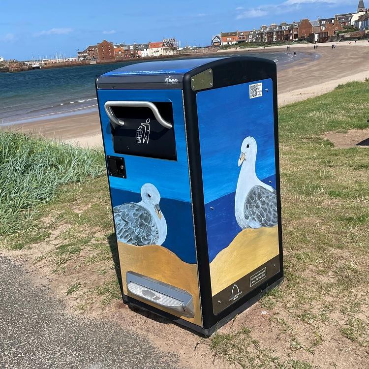A large bin stands beside a path by the sea,  bathed in sunshine, with North Berwick Harbour and West Beach in the background. The bin has a foot peddle at the base for opening the 'trap door' into which waste is discarded. The bin is beautifully and entirely covered in seabird (fulmar) illustrations. A large bin stands beside a path by the sea,  bathed in sunshine, with North Berwick Harbour and West Beach in the background. The bin has a foot peddle at the base for opening the 'trap door' into which waste is discarded. The bin is beautifully and entirely covered in seabird (fulmar) illustrations.